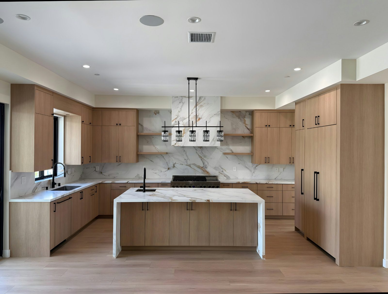 Symmetric high-end kitchen: warm white-oak cabinets, calacatta marble feature wall, integrated appliances, modern linear pendant chandelier, light hardwood floor.