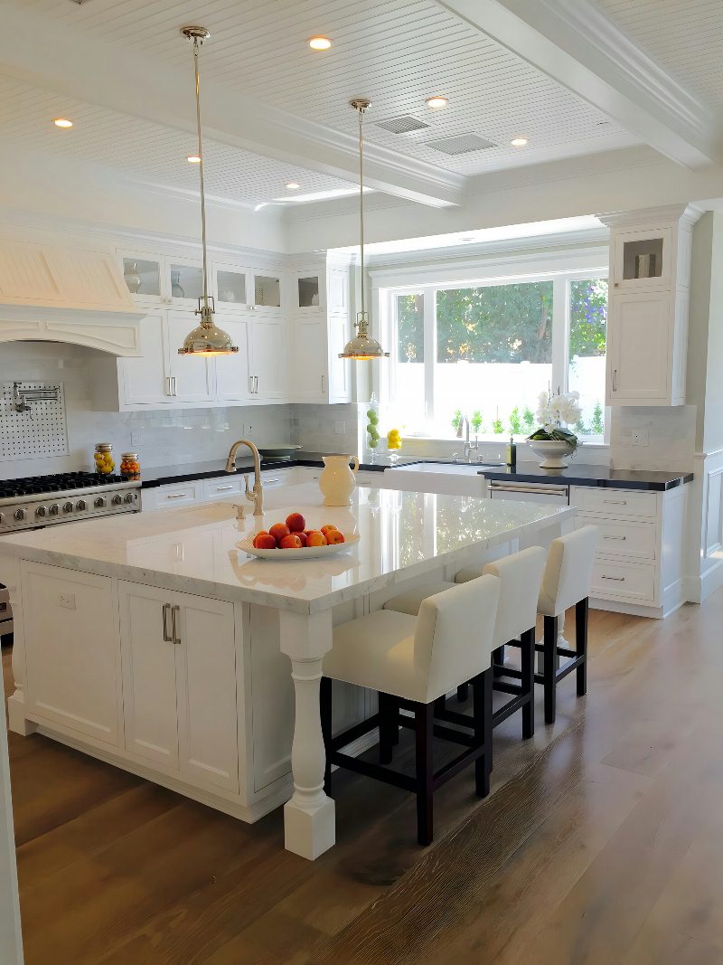 Coastal Cape-Cod kitchen: white shaker cabinetry, turned-leg island, polished nickel pendants, marble counters, Wolf range with red knobs, big window onto greenery.