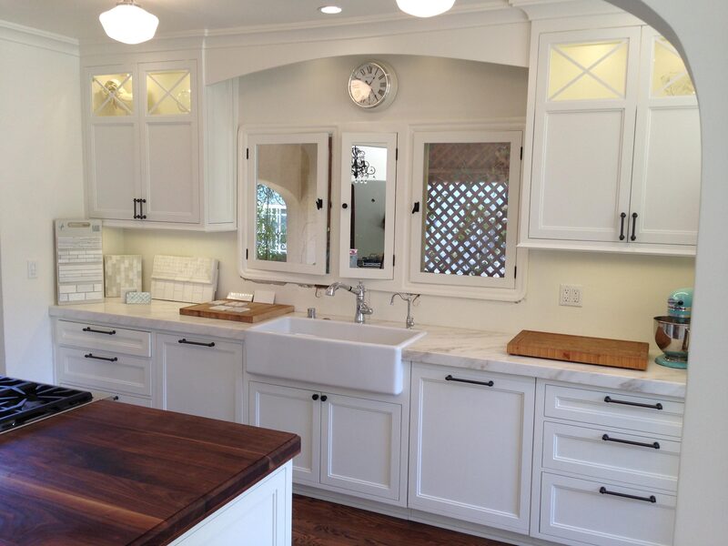 Classic white kitchen: shaker uppers with mullioned glass doors, arched pass-through, farmhouse apron sink, marble counters, butcher block on the island foreground.