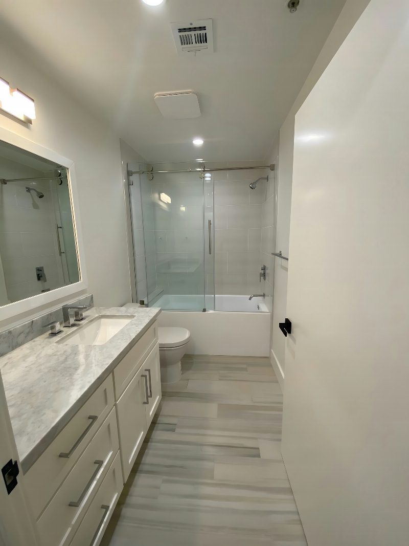Narrow modern bath: white vanity with marble counter, framed mirror, glass shower-over-tub, light wood-plank tile floor.