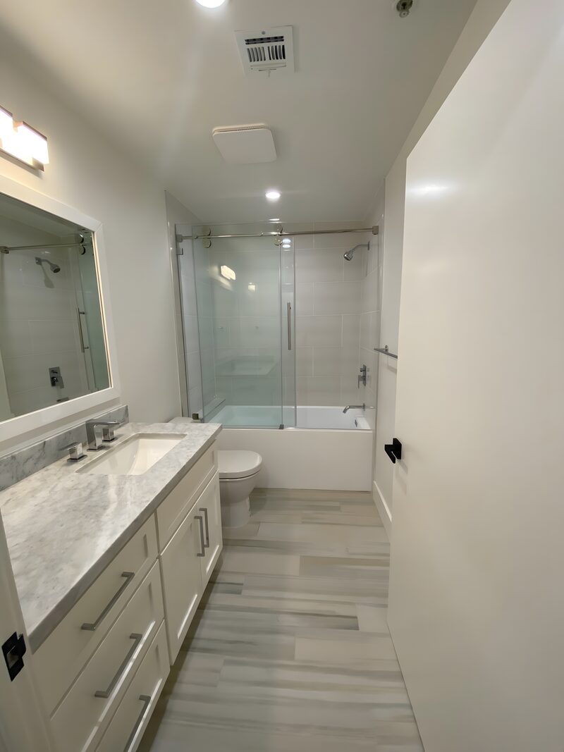 Narrow modern bath: white vanity with marble counter, framed mirror, glass shower-over-tub, light wood-plank tile floor.