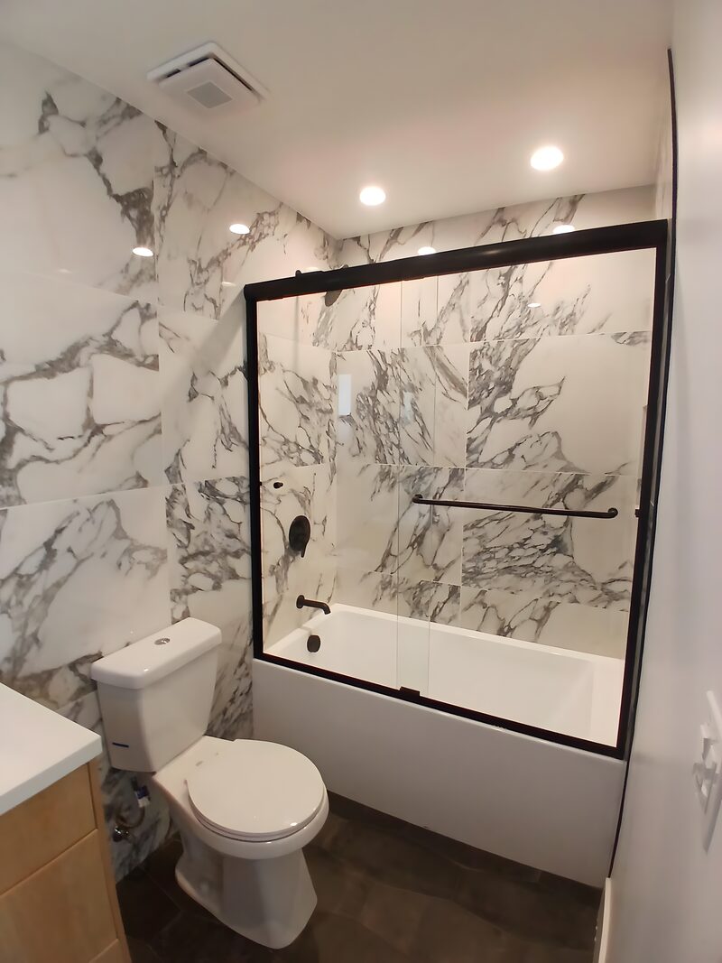 Dramatic bath: full-slab arabescato marble walls, black-framed sliding glass shower over tub, dark stone floor, light wood vanity.