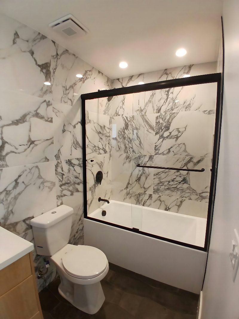 Dramatic bath: full-slab arabescato marble walls, black-framed sliding glass shower over tub, dark stone floor, light wood vanity.